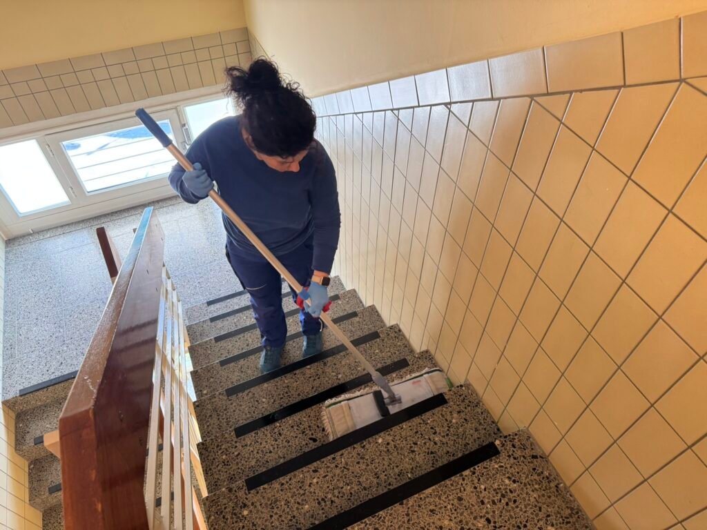 Person in blue gloves mopping a stairwell with tiled beige walls nearby.