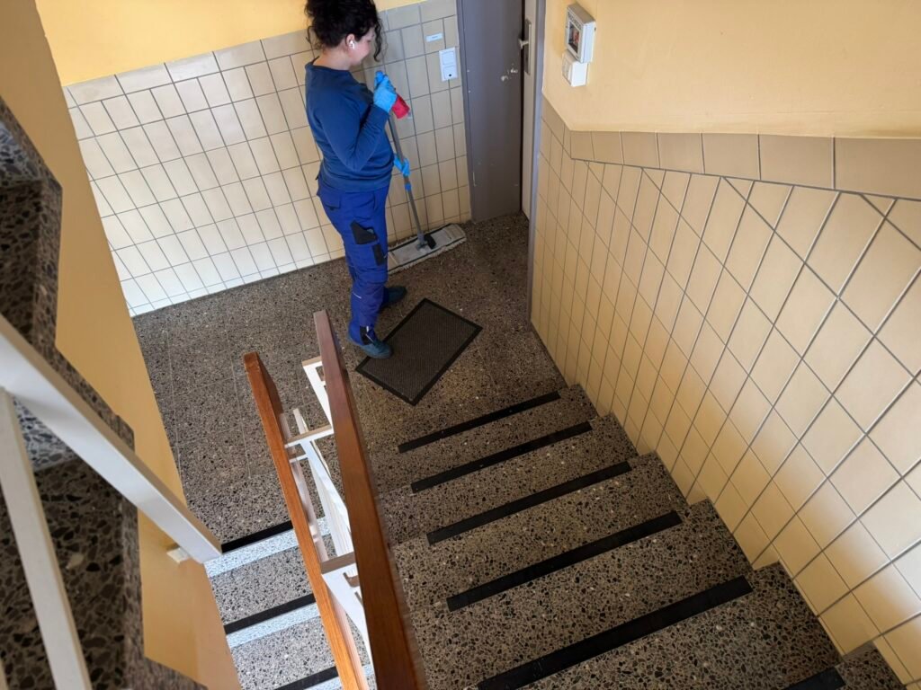 Worker in blue uniform mopping a tiled stairwell with yellow walls, gloves on
