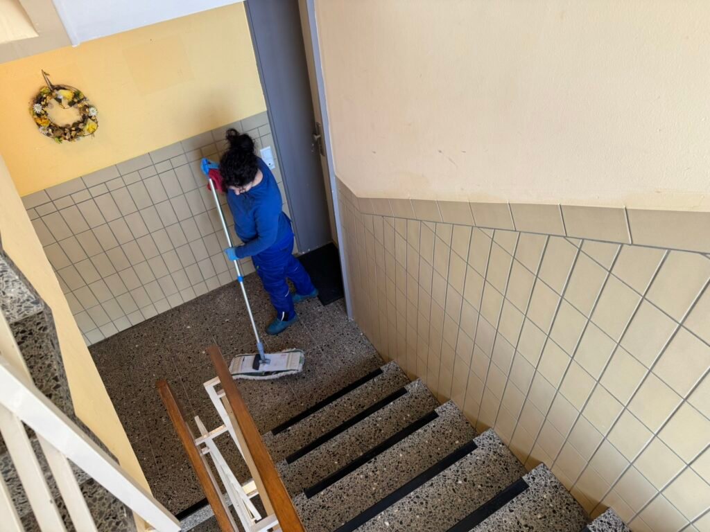 Person in blue cleaning stairs with a mop in a tiled hallway; wreath hanging on yellow wall to the left.